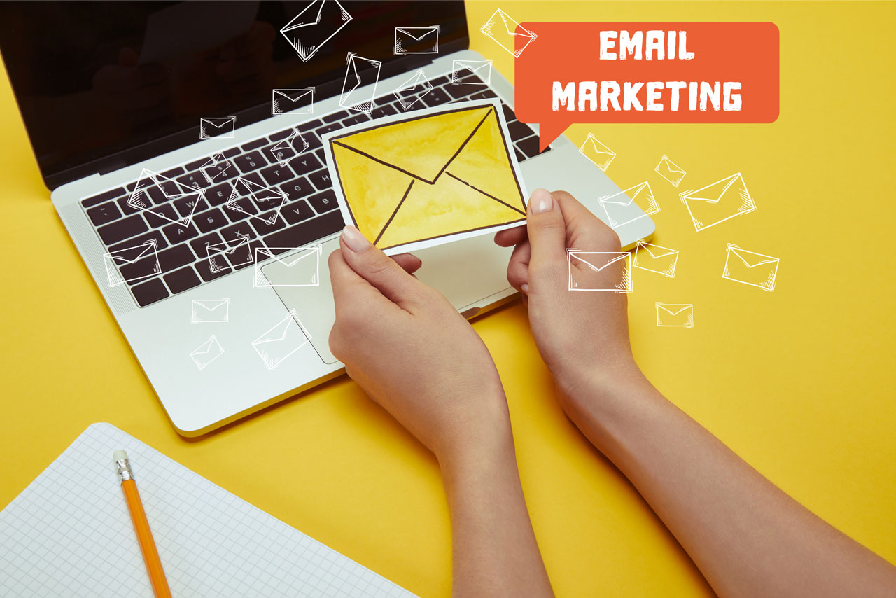 Two hands holding an envelope over a laptop, symbolizing email marketing, set against a vibrant yellow background.